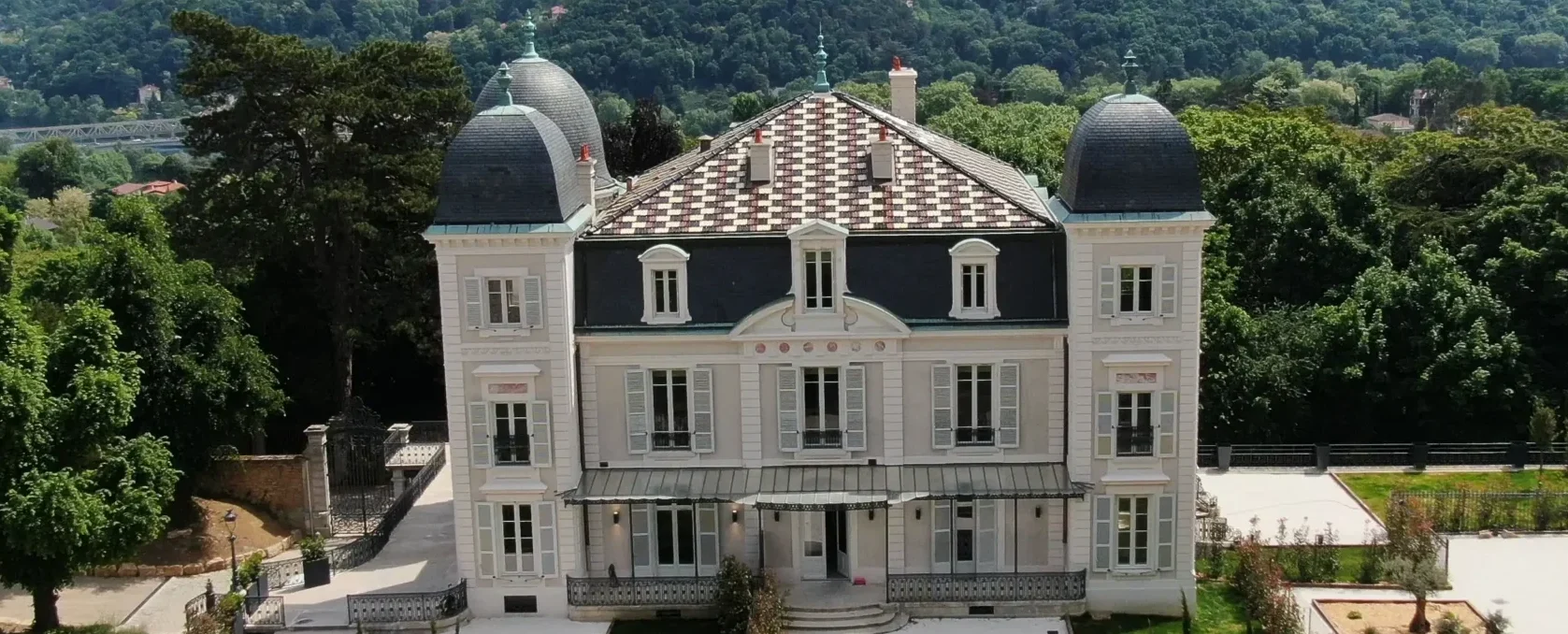 Vue d’une maison historique rénovée à Saint-Cyr-au-Mont-d'Or, avec une toiture en ardoise ornée de motifs géométriques, entourée de verdure et de jardins paysagers.