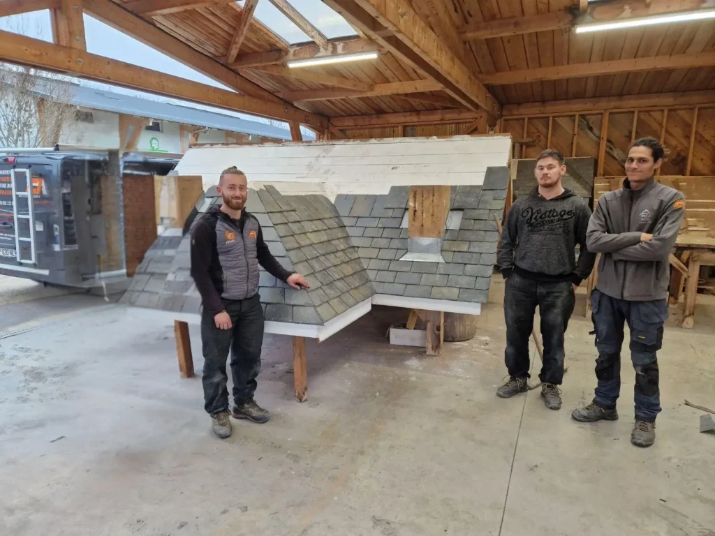 Trois artisans posant devant une maquette de toiture en ardoise réalisée dans un atelier de formation.