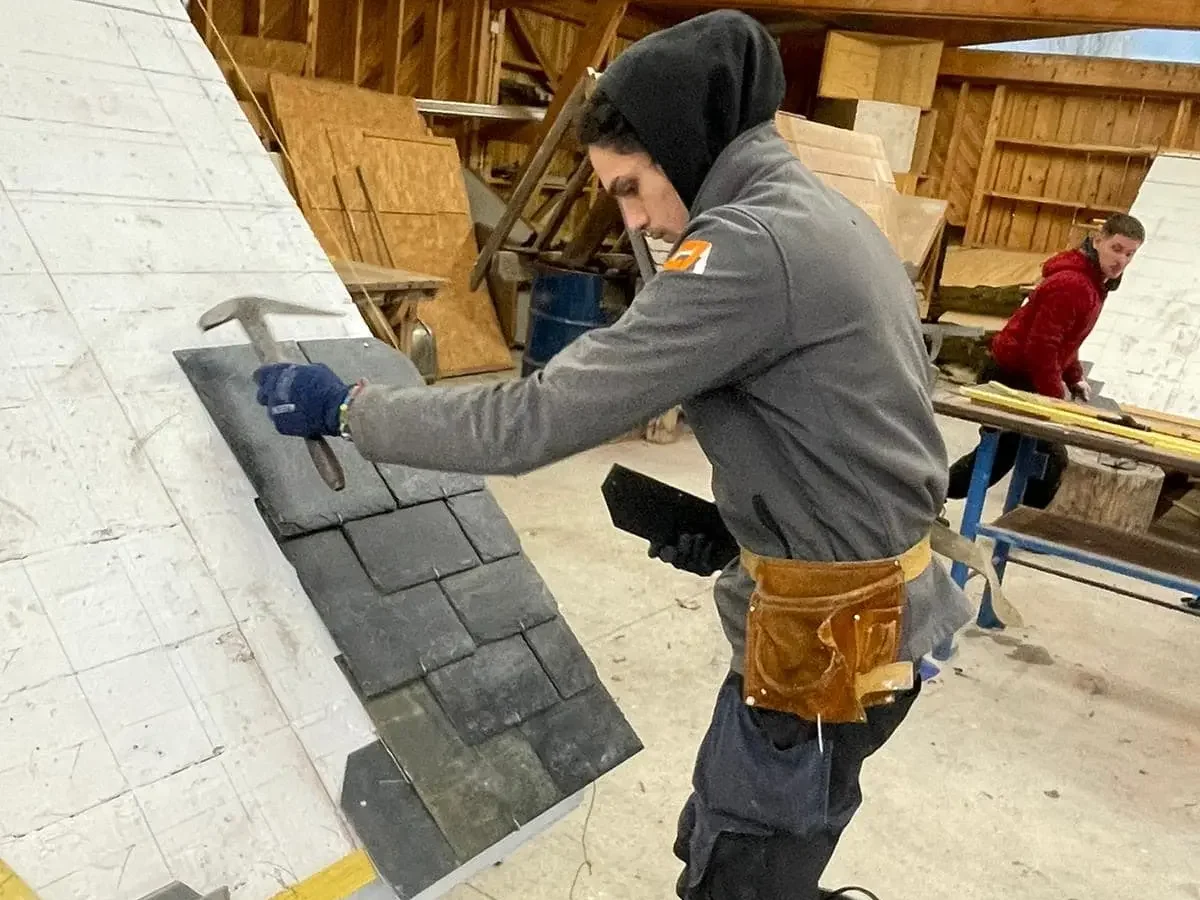 Couvreur posant des ardoises sur une maquette de toiture, utilisant des outils pour ajuster les matériaux dans un atelier.