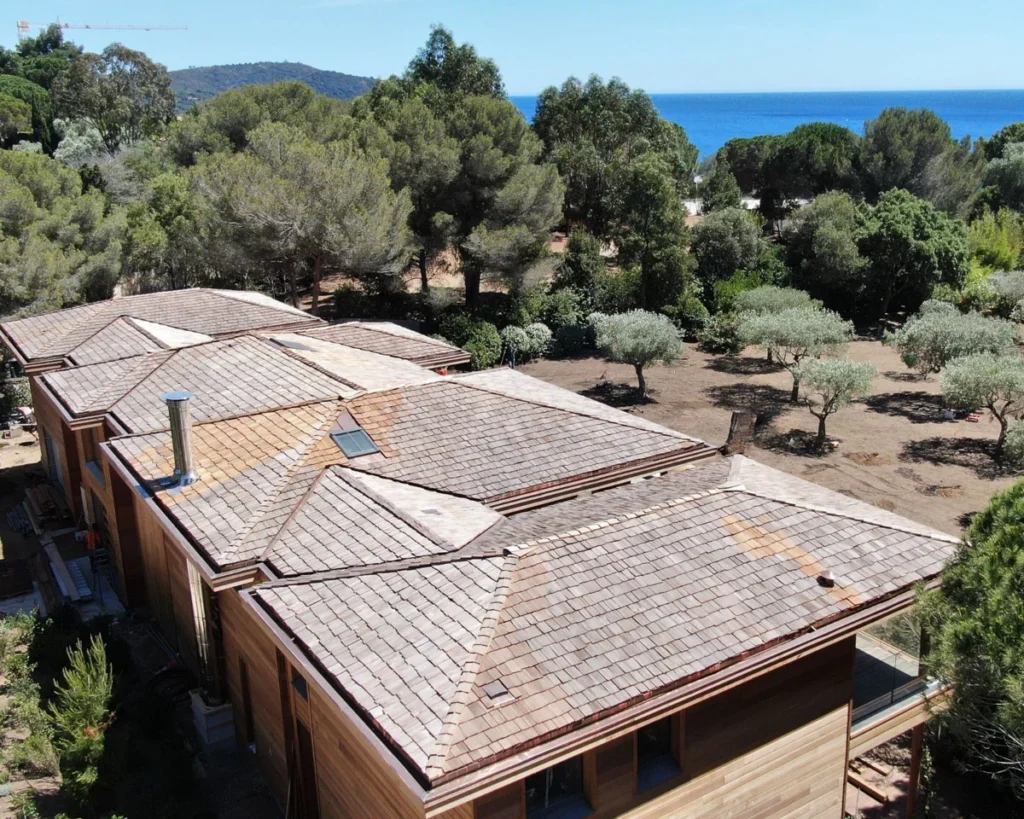 Vue aérienne de toitures en bois au bord de la mer, entourées d'arbres et d'un paysage méditerranéen.