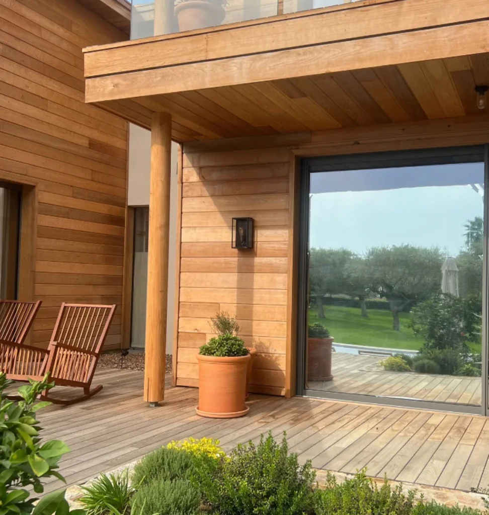 Terrasse en bois avec chaises, pot de fleurs et grande baie vitrée dans un cadre verdoyant.