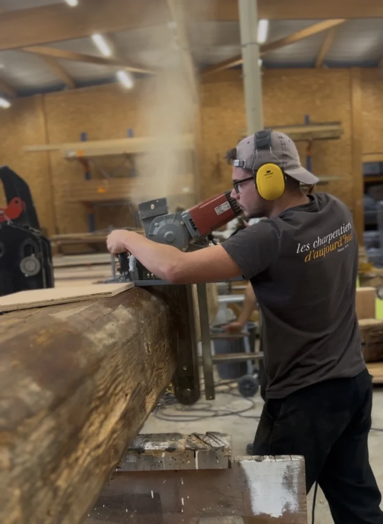 Charpentier en action, taillant une poutre en bois massif dans un atelier.